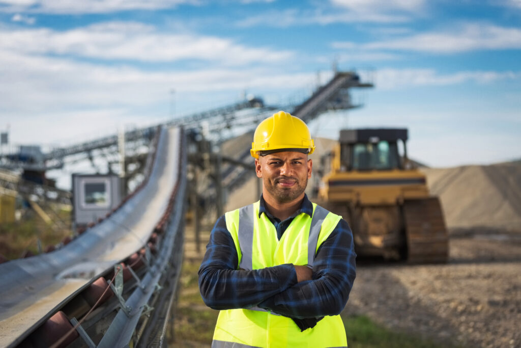Open-pit mine worker wearing protective clothes and yellow helmet standing next to conveyor belt with arms crossed, smiling at camera.